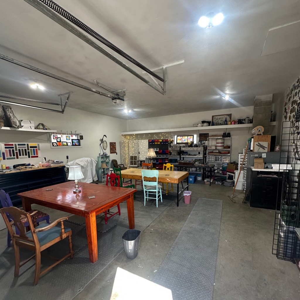 Interior view of a remodeled workshop featuring various tables and chairs, organized shelves with art supplies, and bright lighting.