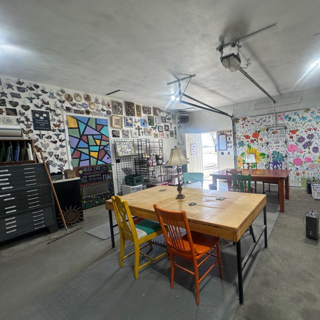 A brightly lit workshop featuring a variety of artwork on the walls, colorful chairs around a large wooden table, and artistic decor.